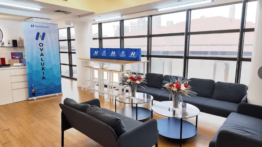 Novaluxia office lounge with grey sofas, round coffee tables and a roller banner beside a window