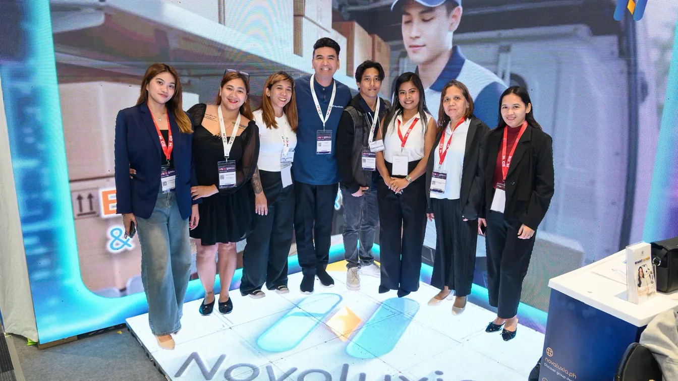 Eight Novaluxia staff in lanyards posing on a branded floor graphic at a retail expo booth