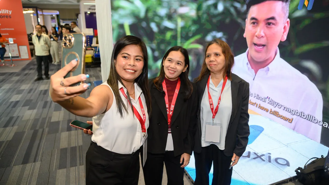 Three visitors taking a selfie in front of a Novaluxia expo booth screen, the kind of photo an is novaluxia legit file marks as the platform's own record